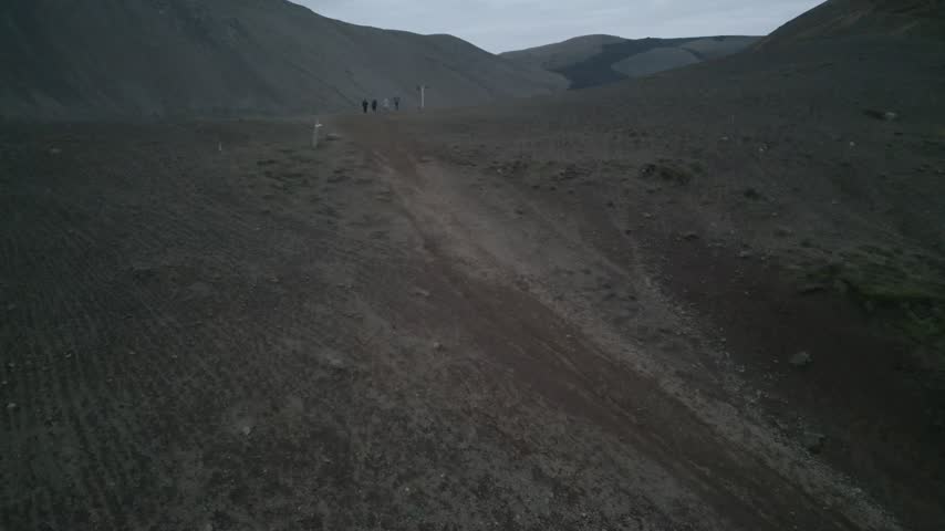 Aerial view of a dirt path winding through the rugged volcanic landscape with people walking in the distance under an overcast sky in Iceland.