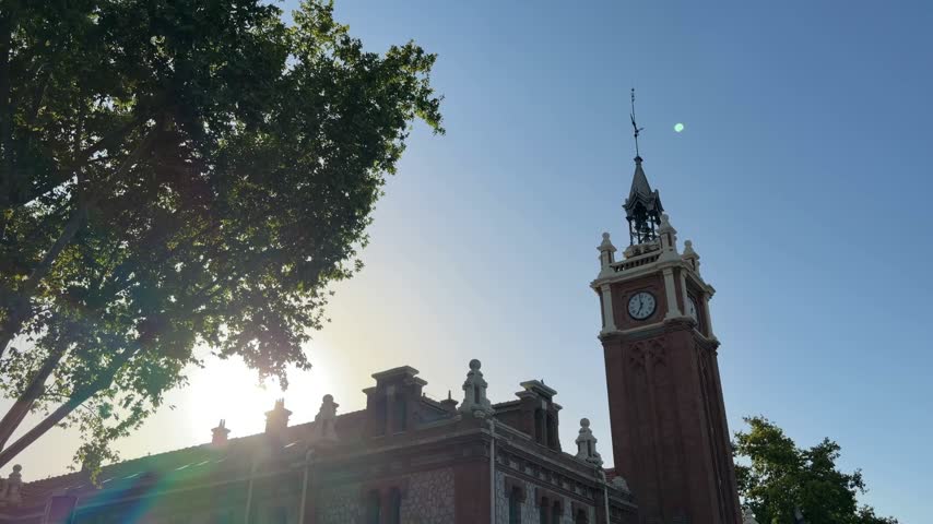 Backlit view of the Casa Consistorial de Arganzuela, a historic clock tower building in Madrid, Spain, with the sun flaring from behind its rooftop and through lush green tree leaves