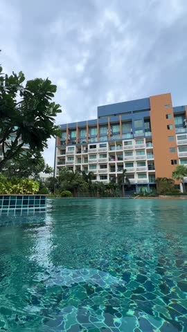 A pool with clear turquoise water at a resort in a residential complex in Pattaya, Thailand. An outdoor pool with a jacuzzi in a condominium surrounded by green trees and flowers. 4К