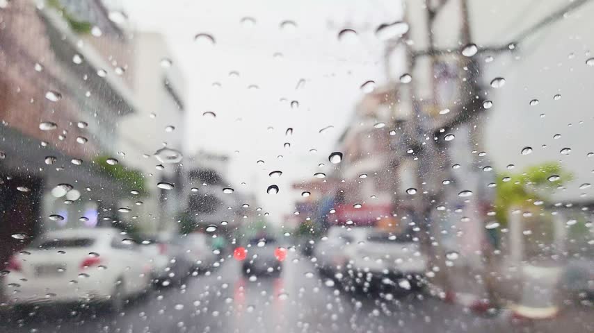 Raindrops on a car windshield with blurred road, vehicles and buildings outside, rainy weather, wet driving conditions, travel and reduced visibility on the road