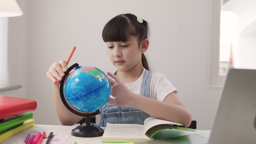Cute Asian schoolgirl sitting at a desk using laptop computer for studying online learning via video calling and a remote teacher while having a geography lesson with a globe model in homeschool room. 