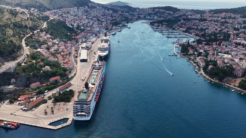 Aerial view of Dubrovnik Bay, Croatia, with tourist and cruise ships sailing through blue Adriatic waters, surrounded by a dramatic coastline, green hills and scenic landscapes.