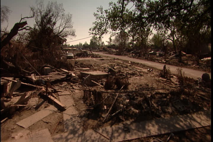 Lot filled with debris, boards and overturned cars, camera pans left to car passing on road in New Orleans after Hurricane Katrina (October 2005).