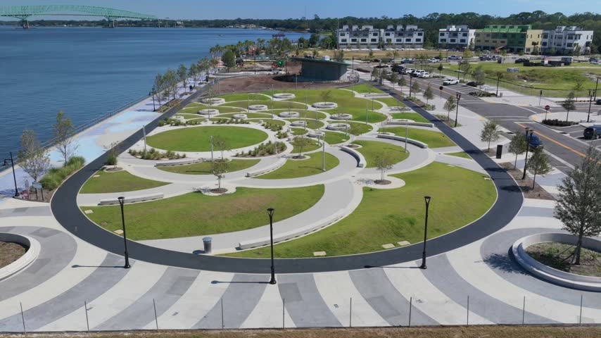 Aerial view of the modern waterfront park with circular pathways, green lawns, and paved walkways along the St. Johns River in Jacksonville, Florida, United States.