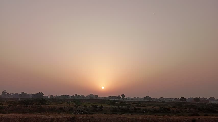 Stunning sunset over a rural village landscape with hazy orange sky, sun setting on the horizon, peaceful evening view with silhouettes of trees and fields.