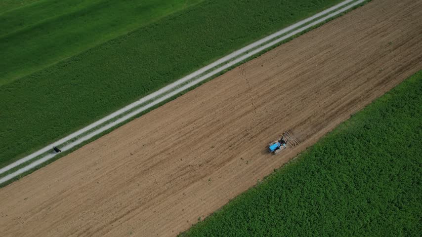Tractor cultivating agricultural field beside green crop and rural road, showing contrasting soil textures and active farming, top-down drone view.