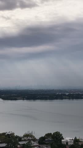 A vertical aerial view drifts over Little Neck Bay as shafts of sunlight break through layered clouds.