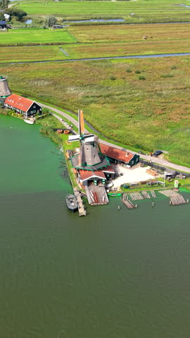 High-angle aerial drone shot capturing a historic Dutch industrial sawmill windmill standing on the waterfront of Zaanse Schans in Zaandam, North Holland, Netherlands. The vertical perspective displays traditional thatched windmill architecture with timber logs floating in the Zaan River basin outside the saw mill slipway, alongside green wooden workshop buildings with red tile roofs. In the background, extensive green polder fields separated by agricultural drainage canals stretch across the flat Dutch countryside under bright summer daylight. High-resolution stock media showcasing Zaanse Schans open-air museum landmarks, authentic 18th-century industrial heritage, North Holland travel attractions, aerial landscape photography, and European cultural tourism. Ideal for vertical social media reels, mobile content creation, travel vlogs, tourism marketing campaigns, educational documentaries, and commercial broadcast media.