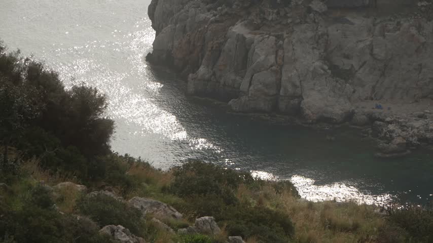 View over Anthony Quinn Bay with a path, rocks and small shrubs in the foreground and a rocky shore with a small pier beyond, Rhodes, Greece