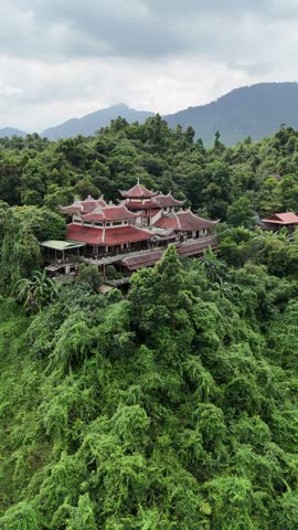 Majestic buddhist temple perched in dense tropical jungle, aerial view revealing lush green mountains and serene lake beneath cloudy sky, traditional vietnamese pagoda landscape in Hue, Vietnam.