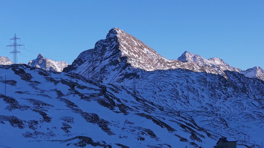 Aerial view of Ospizio Bernina surrounded by snow-covered mountains under a clear blue sky in Poschiavo, Graubunden, Switzerland.
