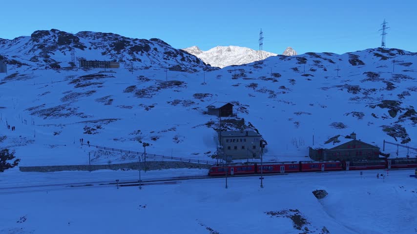 Aerial view of the red Bernina Express train passing by the historic Ospizio Bernina station amidst snow-covered alpine peaks in Poschiavo, Graubunden, Switzerland.
