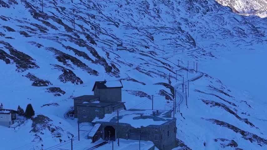 Aerial view of the historic Ospizio Bernina building nestled in a snow-covered mountain landscape with railway tracks winding through the alpine terrain in Poschiavo, Graubunden, Switzerland.