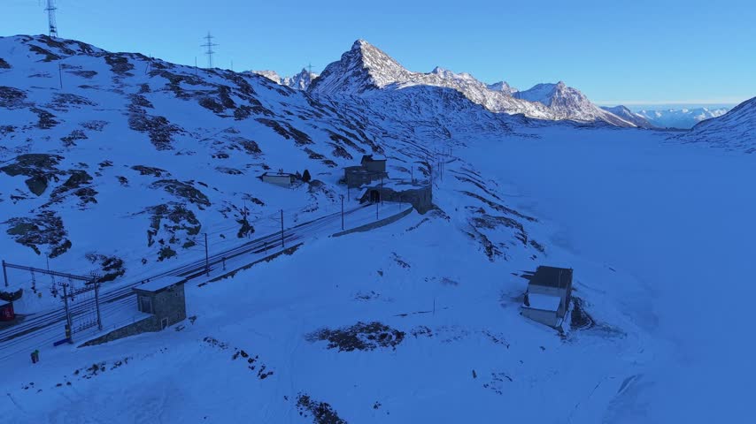 Aerial view of Ospizio Bernina surrounded by snow-covered mountains, railway tracks, and stone buildings under a clear blue sky in Poschiavo, Graubunden, Switzerland.
