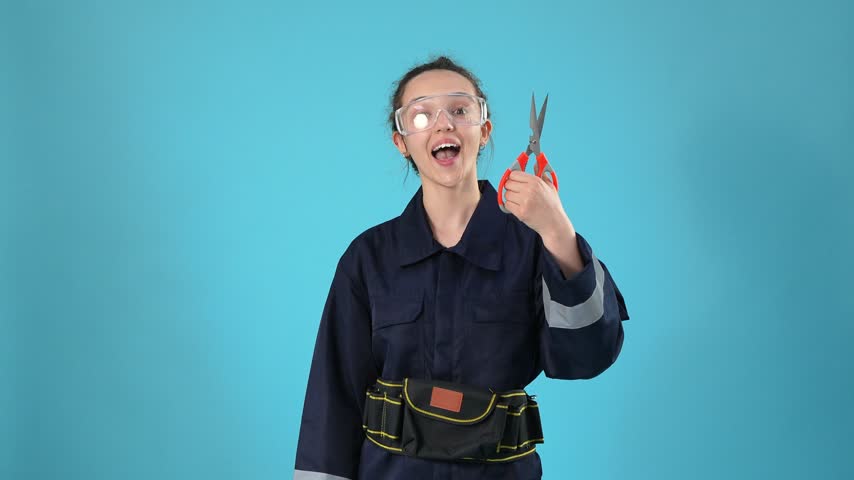 A young female worker wearing a dark jumpsuit, safety glasses, and a tool belt holds red scissors and opens them while smiling against a bright blue studio background.