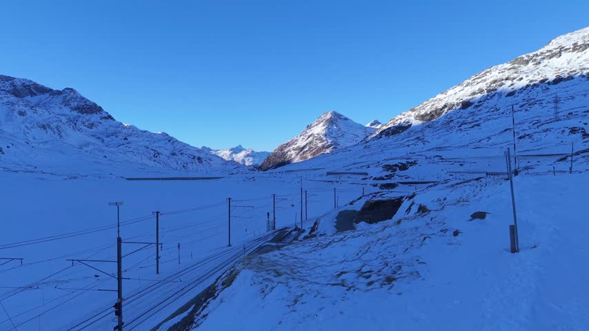 Aerial view of Ospizio Bernina with snow-covered mountain landscape and railway tracks under a clear blue sky in Poschiavo, Graubunden, Switzerland.