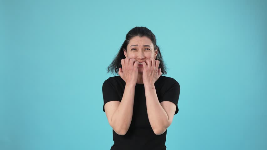 A frightened woman bites her nails and trembles with anxiety against a bright cyan background. She looks around nervously with a worried expression.