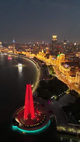 Aerial view of the iconic Bund waterfront in Shanghai, China at night, showcasing the illuminated historical buildings, bustling promenade, and boats cruising along the Huangpu River from above.