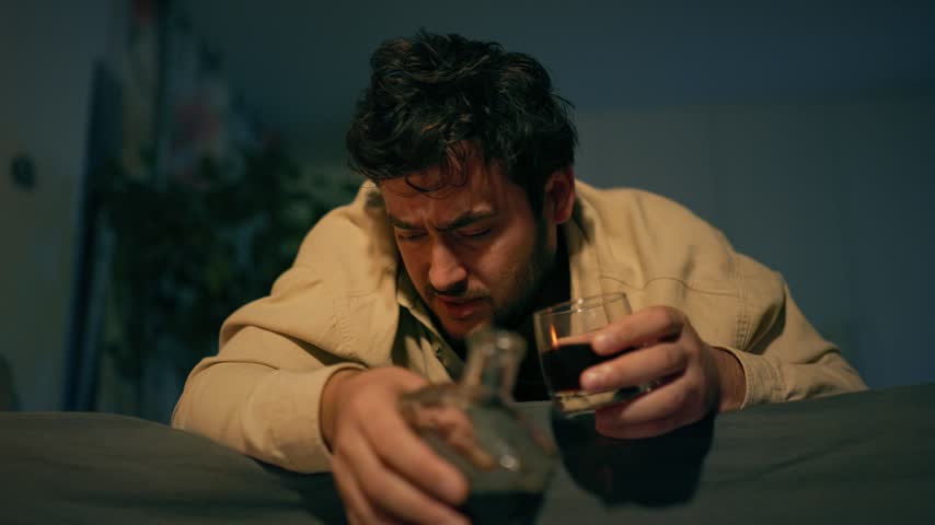 Experiencing a blackout from alcohol abuse, an adult male lies completely motionless face down on his bed, illuminated by moody bedroom lighting.