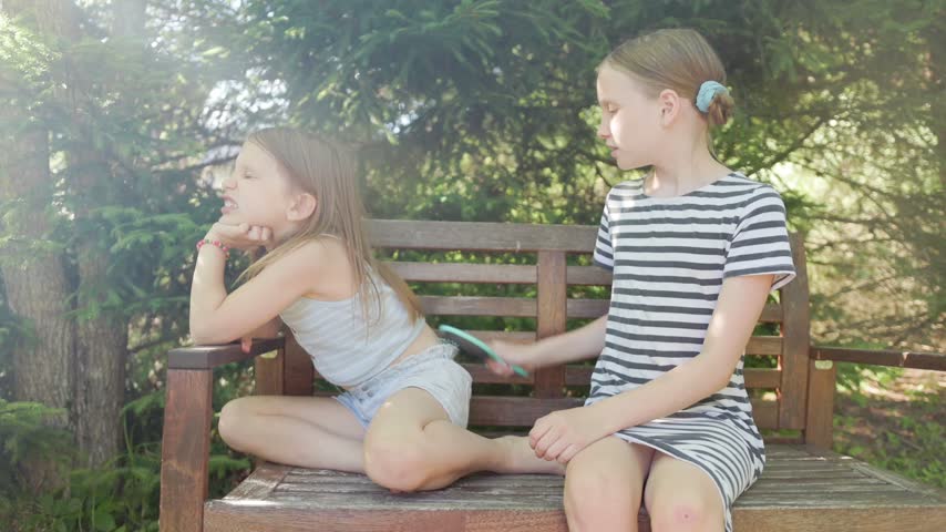 Young girl rests peacefully while sister carefully grooms long hair using soft brush on garden bench. Sisters share affectionate bonding moment amidst lush evergreen trees in bright sunlight.