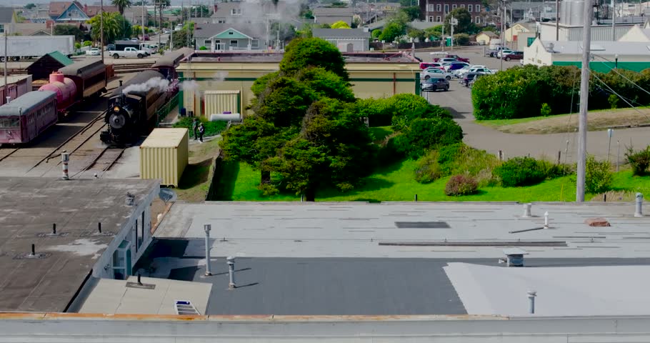 Aerial view of a historic steam train moving along tracks near a depot, surrounded by town buildings, green trees, and parking areas in Fort Bragg, California, United States.