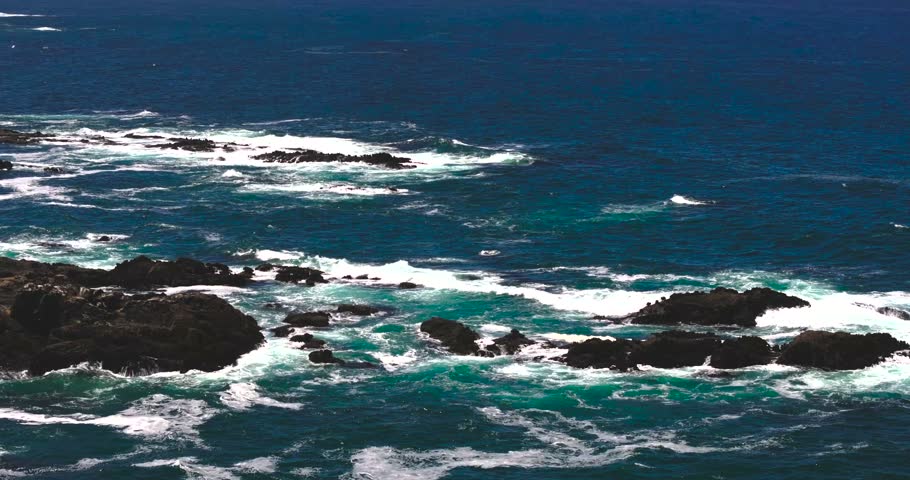 Aerial view of Pacific Ocean waves crashing against jagged dark rocks along the rugged coastline in Fort Bragg, California, United States.