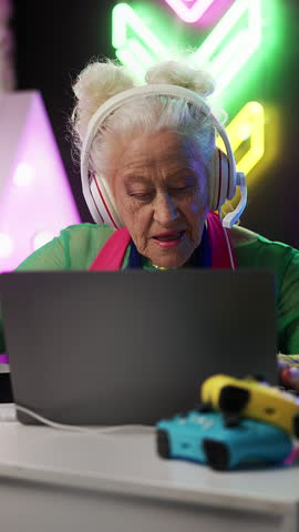 Stylish granny in a headset playing a video game on laptop, celebrating victory