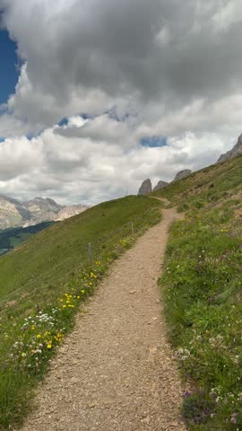 Dirt hiking trail climbing through green alpine meadows toward distant peaks in the Dolomites, Italy, under big clouds and blue sky