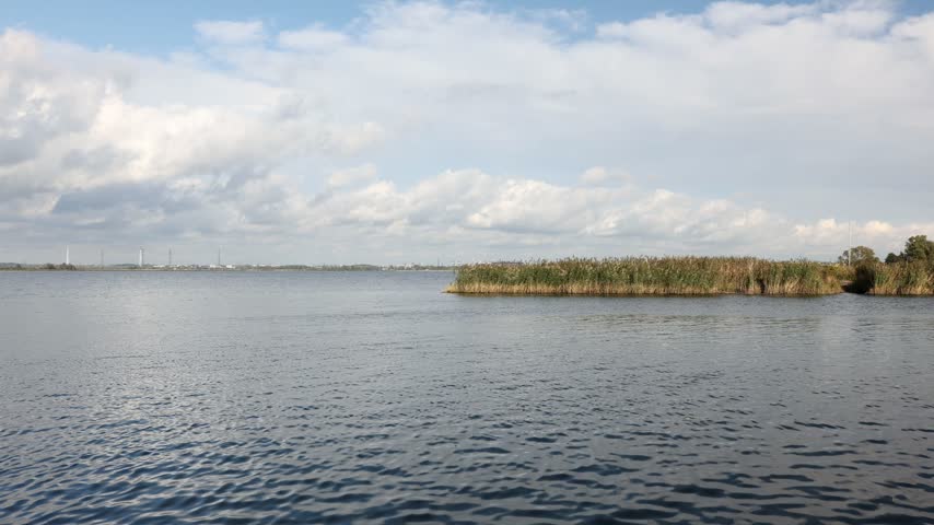 Calm lake with rippling water, reeds and cloudy blue sky on sunny day, natural lakeside landscape