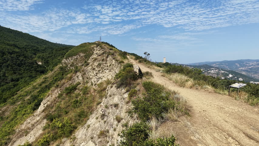 Dirt hiking trail on mountain ridge above lake in summerDescription: Scenic dirt pathway along a rocky mountain ridge overlooking a peaceful lake, dense green forests, and surrounding hills under a bright sky with light clouds.