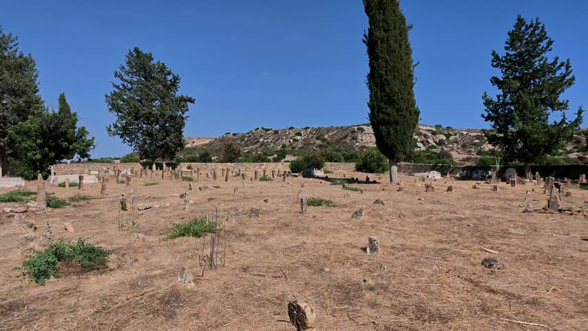 Tochni Turkish Cypriot cemetery in Cyprus with numerous old stone grave markers across dry ground, tall cypress and pine trees, rocky hills and a clear blue sky