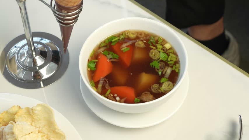 Hot vegetable soup with carrots potatoes and herbs in white bowl on restaurant table