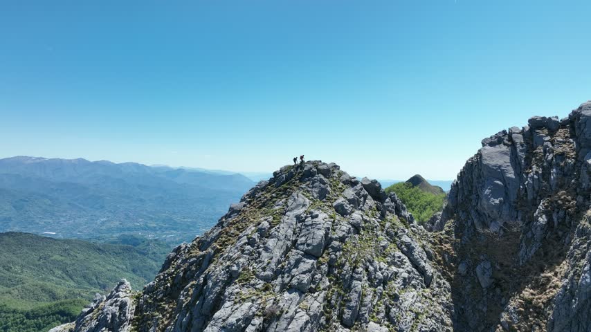 Aerial view of Pania Secca mountain peak with two hikers standing on the rocky summit under a clear blue sky in Vagli Sotto, Tuscany, Italy.
