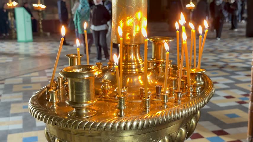 Close up shot of thin beeswax candles burning on golden brass candlestick inside a church as people walk in the background. Russian orthodox cathedral interior, religious prayer devotion ritual.