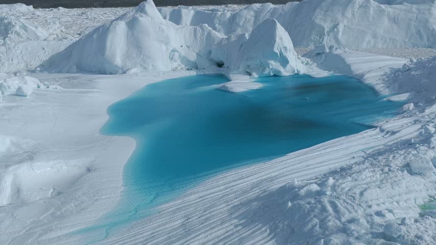 Aerial view of Ilulissat Icefjord featuring a vibrant turquoise meltwater pool surrounded by massive white icebergs under bright daylight in Ilulissat, Avannaata, Greenland.