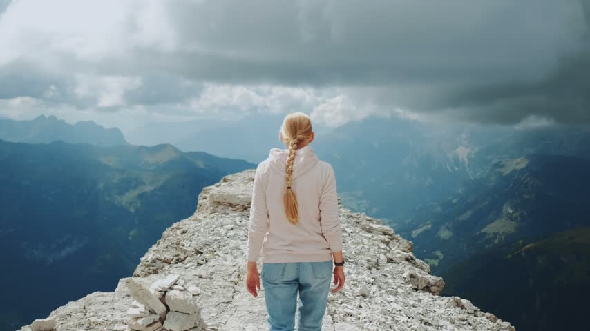 A blonde woman with a braid walks on a rocky mountain peak surrounded by vast mountain ranges and dramatic cloudy sky during daytime.