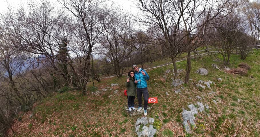 Couple on trail through bare trees on hillside of Monte Barro with mountains in background in Italy. Aerial backward