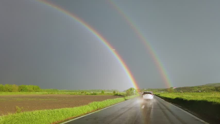 Scenic view from car driving along country road with vibrant double rainbow spanning across dark stormy sky after spring rain.