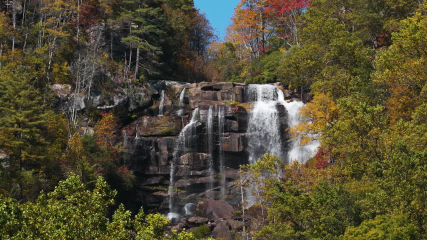 Scenic view of Whitewater Falls in fall season. Cascading water tumbles over rugged rocks amid vibrant yellow and orange forest trees.