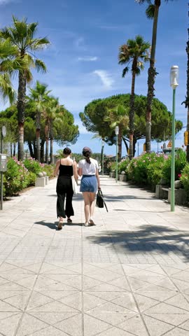 Rear view of two young women strolling side by side on stone promenade surrounded by palm trees and blooming flowers on sunny summer day.