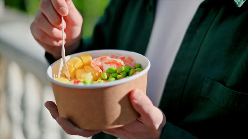 Close up of person enjoying healthy shrimp poke bowl lunch on sunny day outside with wooden spoon.