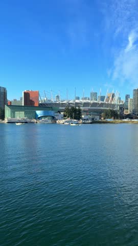 A sailboat glides across False Creek with the downtown Vancouver skyline, BC Place stadium and condo towers behind it under a bright blue sky in winter.