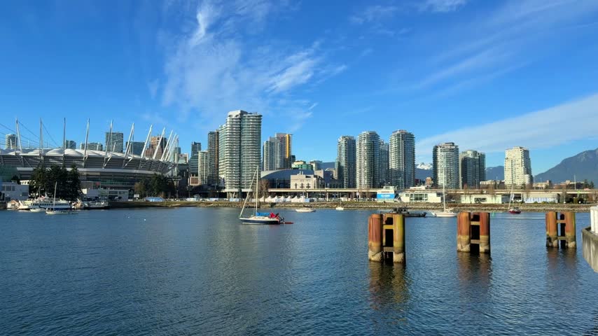 A sailboat glides across False Creek with the downtown Vancouver skyline, BC Place stadium and condo towers behind it under a bright blue sky in winter.