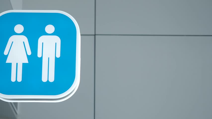Close up shot showing blue square toilet sign with white gender symbols mounted on white wall inside clean office or shopping mall hallway indicating public restroom location.