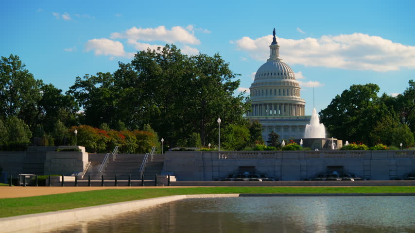Locked off wide shot of the US Capitol dome, fountain and grounds behind a reflecting pool in Washington DC as white clouds drift across a blue sky.
