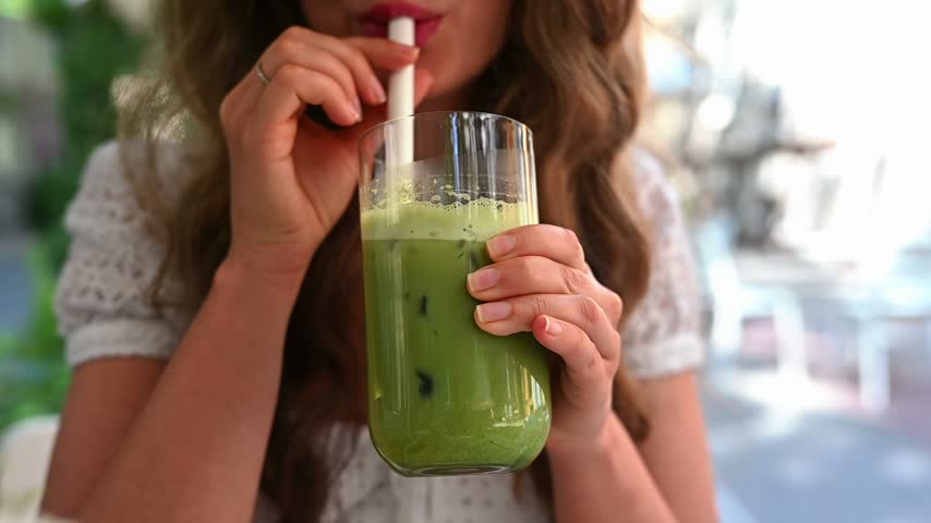 Young woman enjoying cold green tea beverage holding glass with ice while sitting at table in sidewalk coffee shop terrace.