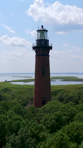 Iconic Red Brick Currituck Beach Lighthouse Stands Above Coastal Forest Overlooking Calm Waters In Corolla, North Carolina, United States. - aerial vertical shot