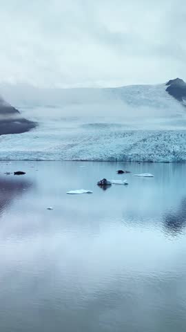 Aerial view of calm glacial lagoon with blue ice chunks floating on water surface against dramatic foggy mountain glacier.