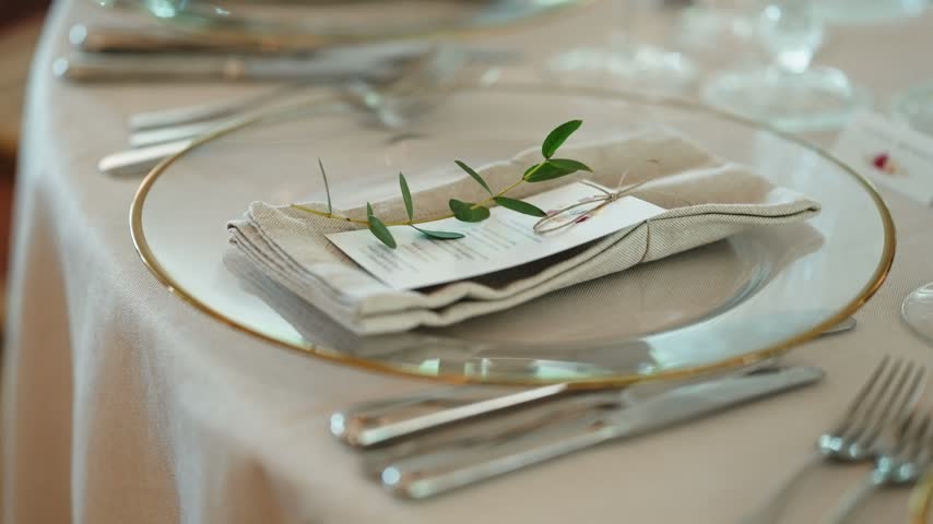 Gold rimmed glass charger holds folded linen napkin and menu card tied in twine with a green sprig, cutlery set around it.