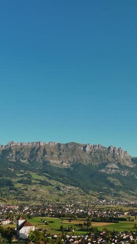 Vertical aerial view over Werdenberg, Switzerland, showing its historic lakeside settlement and green Rhine Valley farmland beneath the dramatic Churfirsten mountain range.